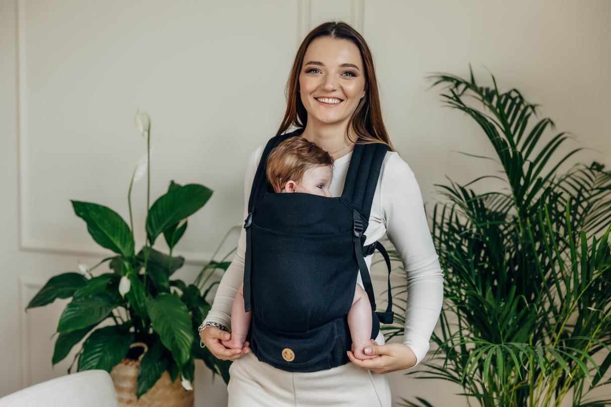 Mom wearing black cotton LennyLight baby carrier with infant, surrounded by green plants.