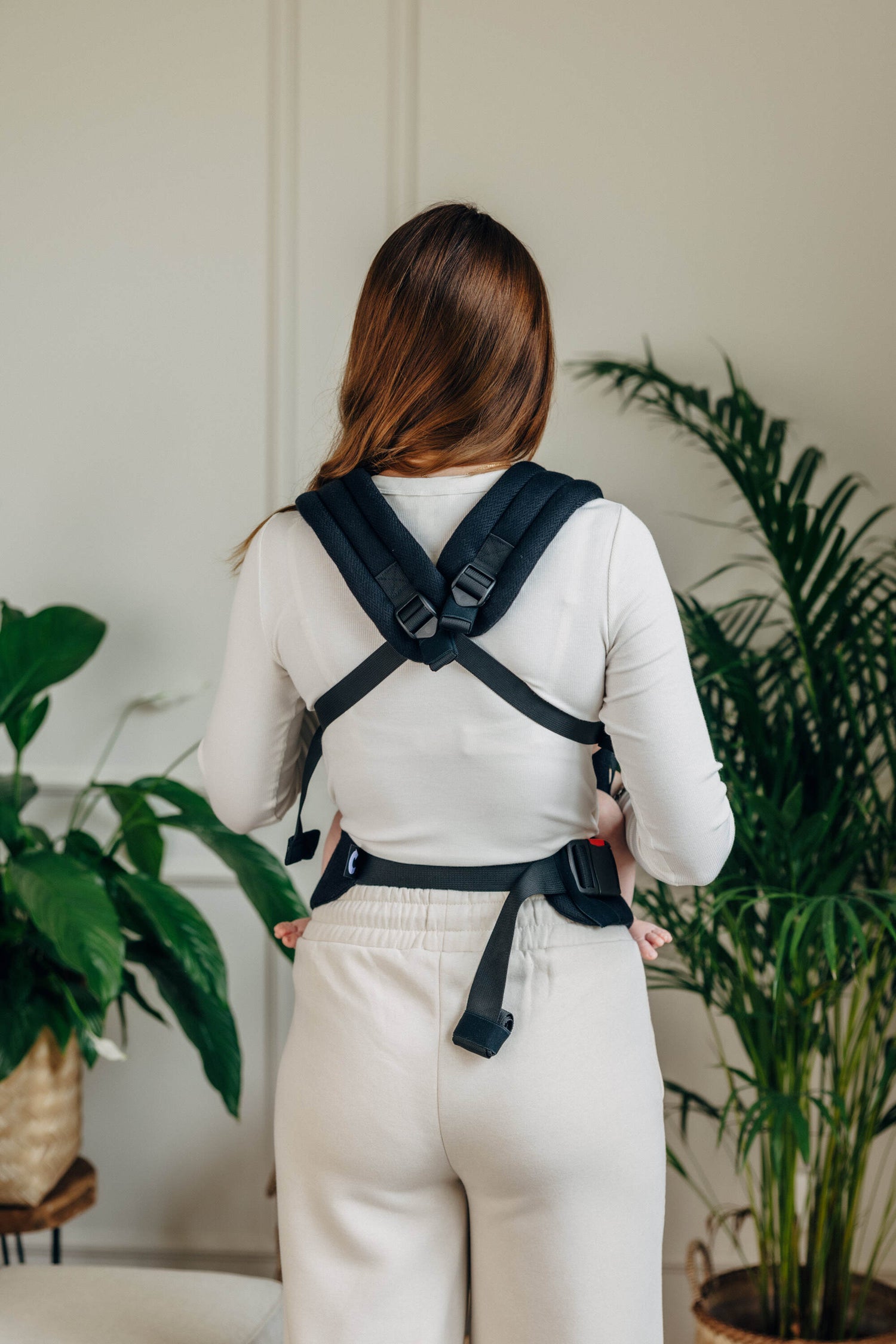 Back view of a woman wearing the LennyLight black cotton baby carrier in a cozy indoor setting.