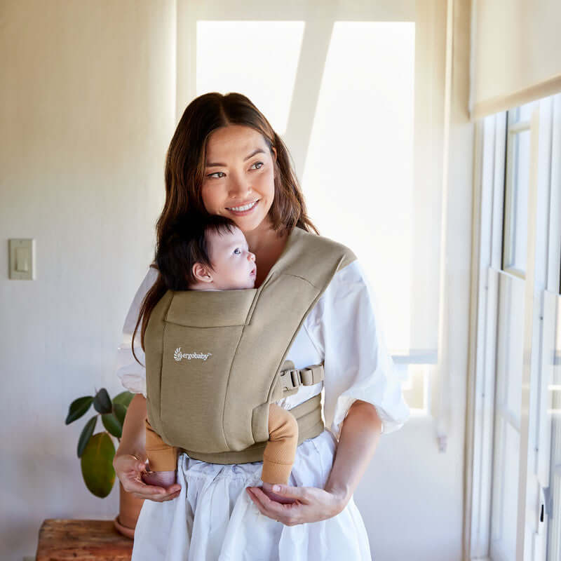 Mom wearing Ergobaby Embrace - Soft Olive baby carrier with smiling baby indoors | Babymaxi
