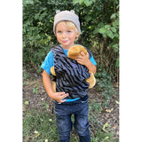 Child wearing Manduca Doll Carrier - Zebra, carrying a teddy bear in front of them, in a natural outdoor setting | Babymaxi